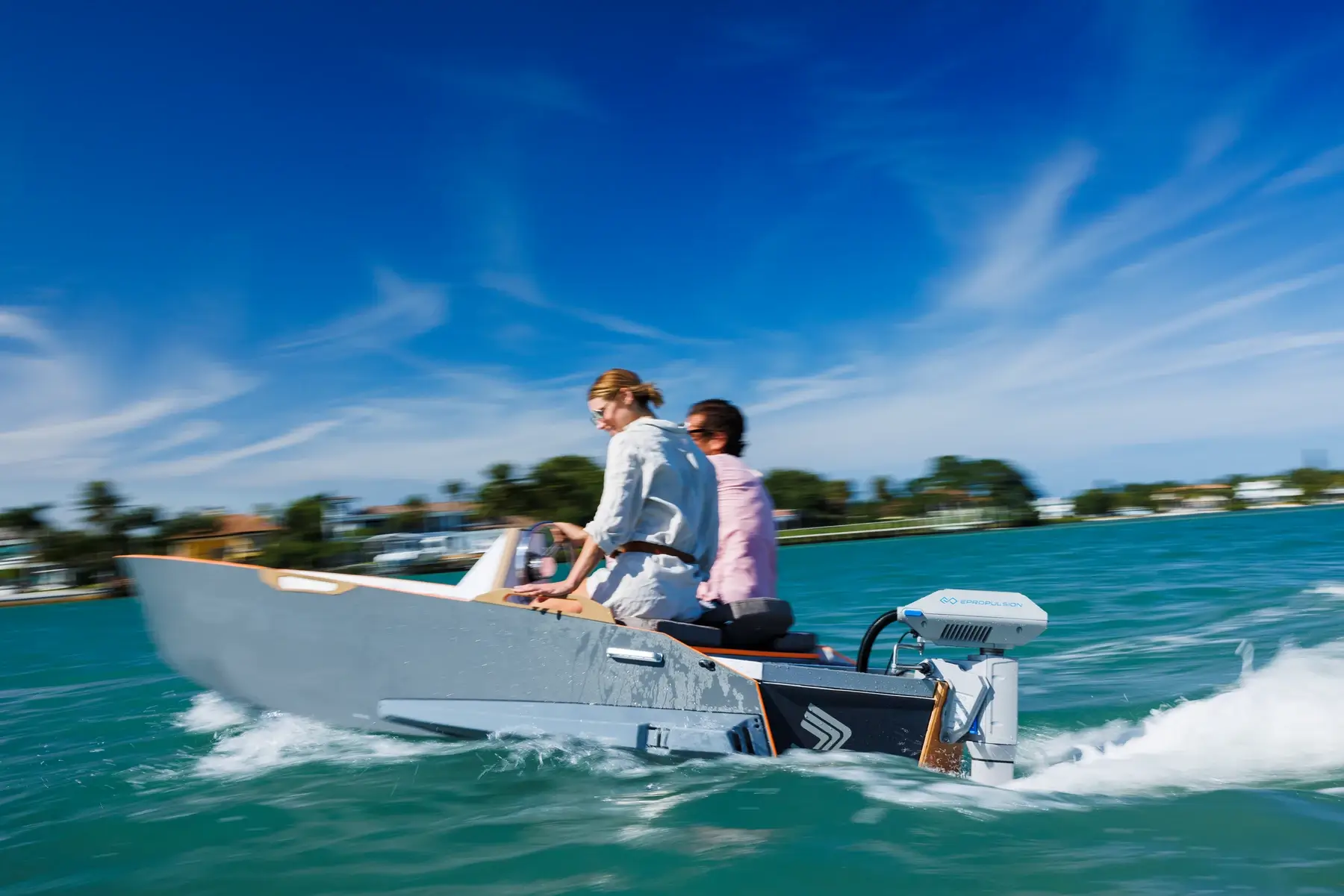 Two individuals ride in a sleek electric boat powered by an ePropulsion electric motor on a sunny day. The boat speeds across green-blue water with trees and buildings visible in the blurred background, emphasizing its movement. The sky is clear with a few wispy clouds. Oceans EV