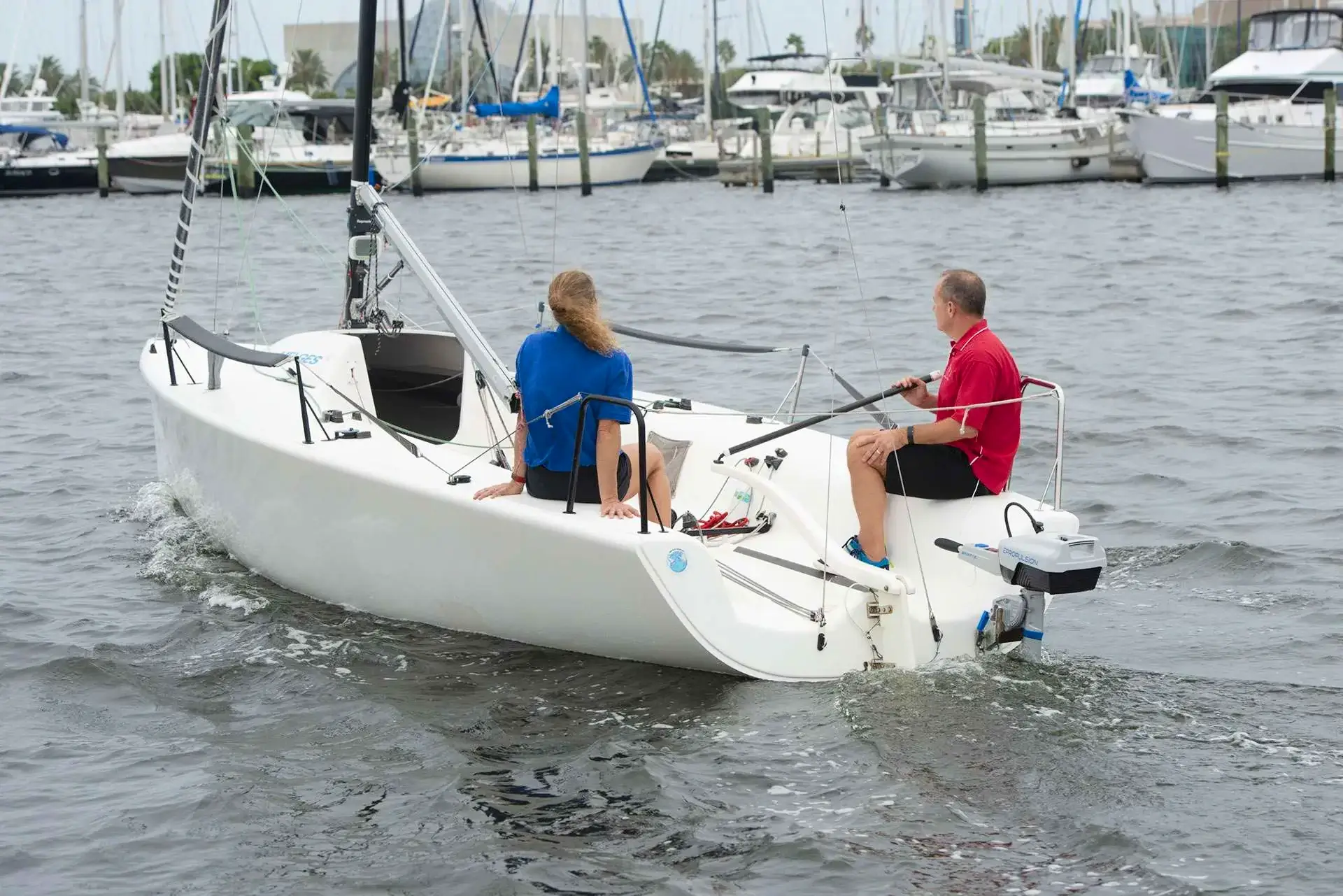 Two people are sailing a small white boat with an ePropulsion electric motor on a calm marina, surrounded by various larger boats docked in the background. One person is wearing a blue shirt and the other a red shirt, both managing the boat's tiller and main sheet. Oceans EV